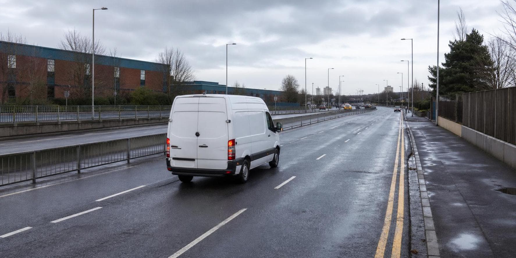 Plain white courier van travelling on a wet urban road in Enfield with commercial buildings beyond